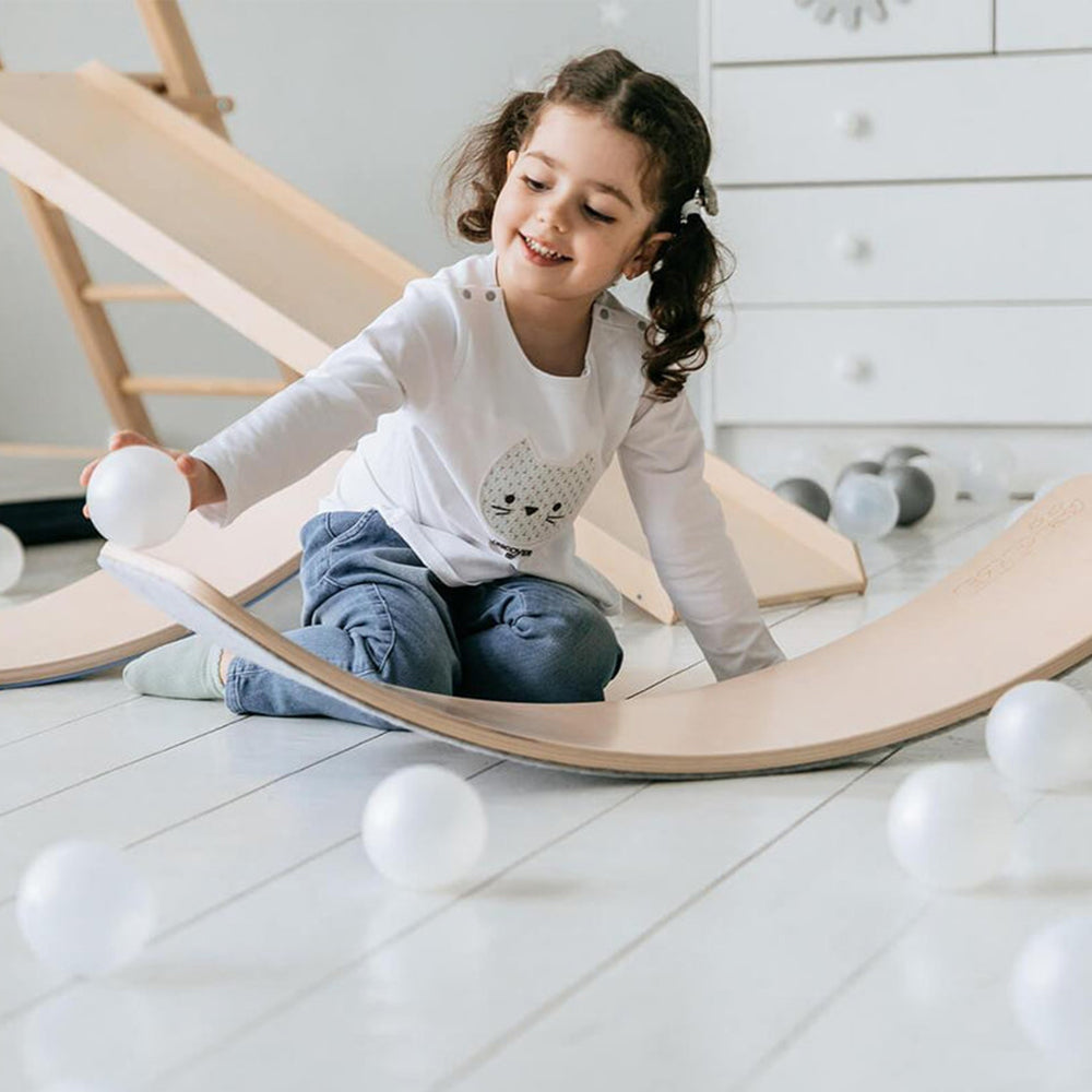 A girl smiling and playing with a natural wooden balance board surrounded by soft balls