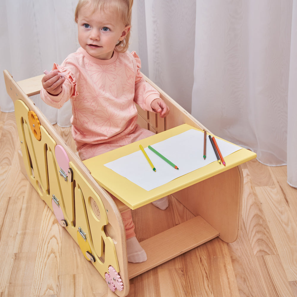 Montessori learning tower made of natural wood with a yellow side busy board, featuring interactive gears, sliders, and lockable elements for toddlers.