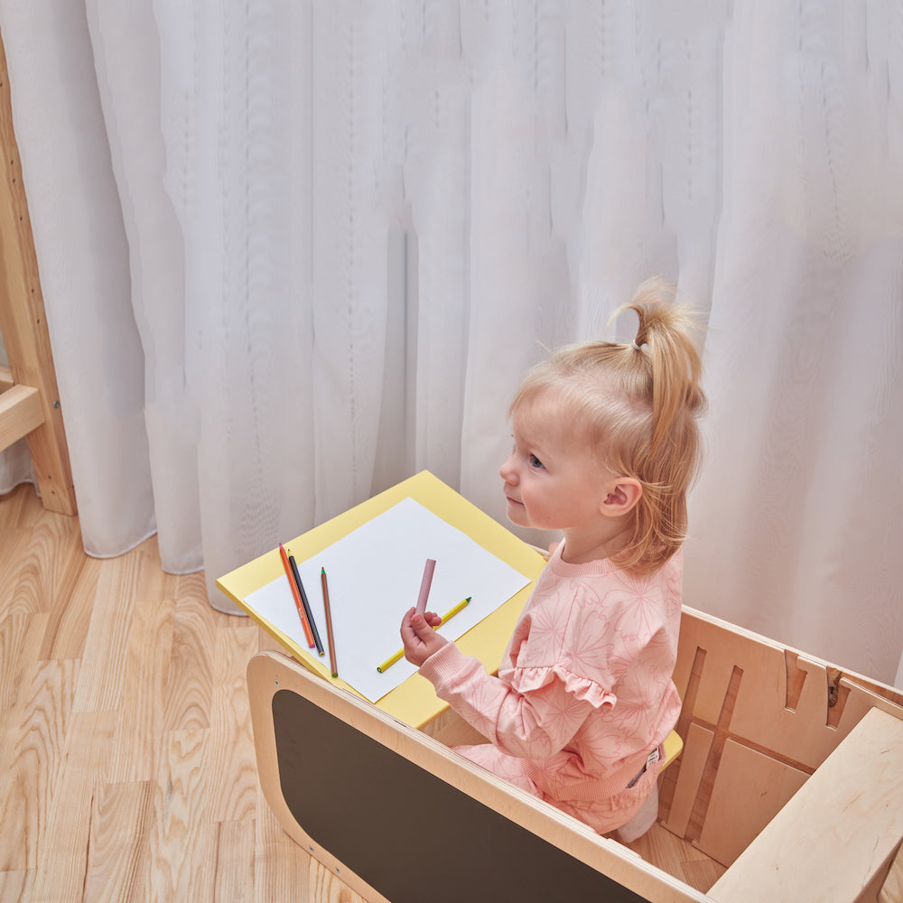 Montessori learning tower made of natural wood with a yellow side busy board, featuring interactive gears, sliders, and lockable elements for toddlers.