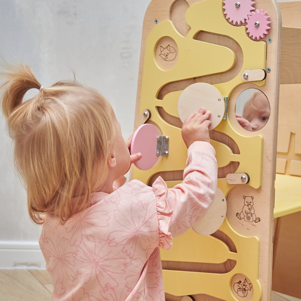 Montessori learning tower made of natural wood with a yellow side busy board, featuring interactive gears, sliders, and lockable elements for toddlers.