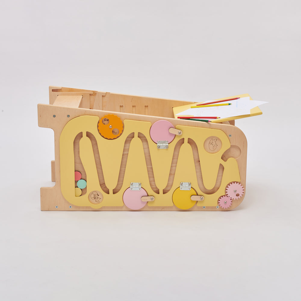 Montessori learning tower made of natural wood with a yellow side busy board, featuring interactive gears, sliders, and lockable elements for toddlers.