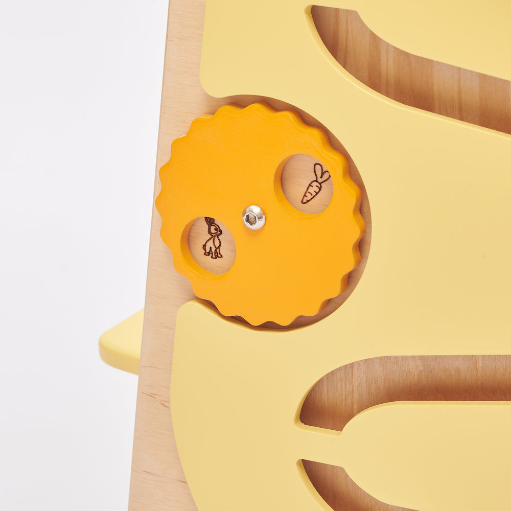 Montessori learning tower made of natural wood with a yellow side busy board, featuring interactive gears, sliders, and lockable elements for toddlers.