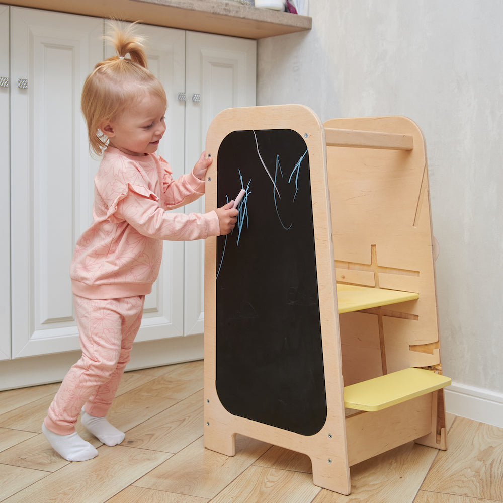 Montessori learning tower made of natural wood with a yellow side busy board, featuring interactive gears, sliders, and lockable elements for toddlers.