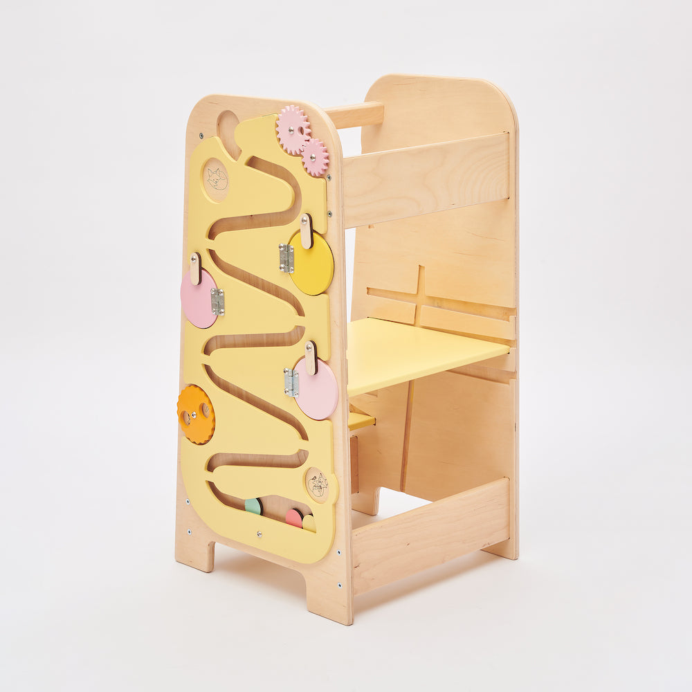 Montessori learning tower made of natural wood with a yellow side busy board, featuring interactive gears, sliders, and lockable elements for toddlers.