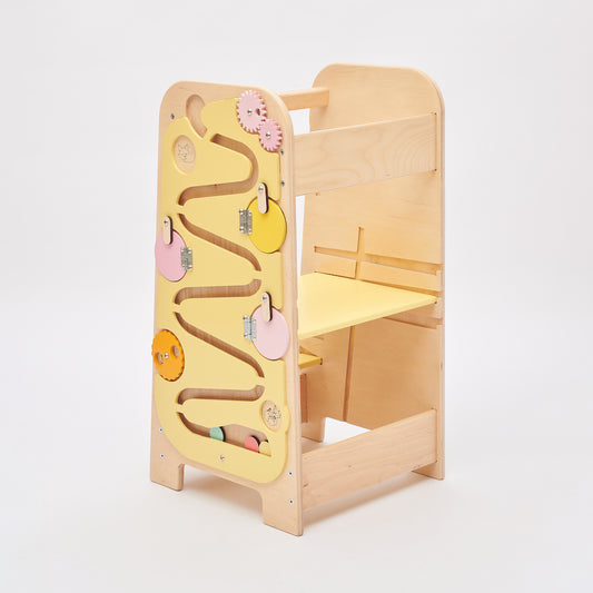 Montessori learning tower made of natural wood with a yellow side busy board, featuring interactive gears, sliders, and lockable elements for toddlers.